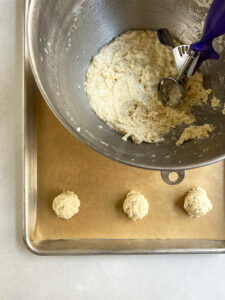 Three scoops of macaroon batter on a parchment-lined baking sheet with the bowl of batter and the scoop nearby.