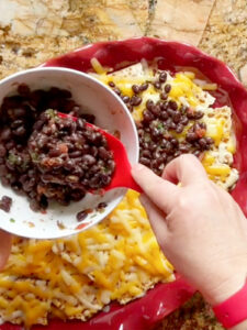 Black beans in a white bowl being spooned on matzo nachos with a red spoon.