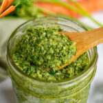 Pinterest image for pesto with carrot tops, parsley, arugula, macadamia nuts and preserved lemon in a jar with a wooden spoon.