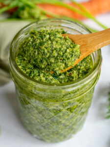 Carrot top pesto in a jar with a close up on a wooden spoon with a dollop of pesto on it.