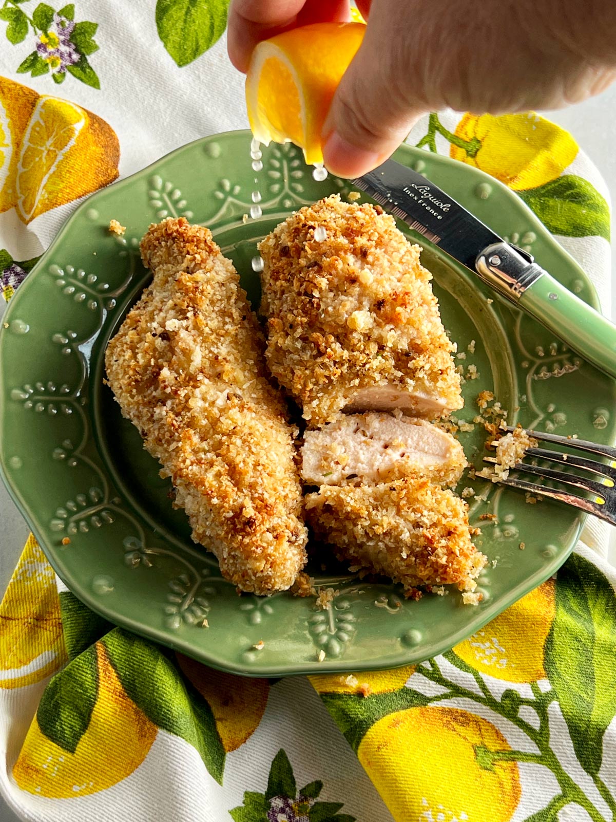Baked breaded lemon chicken on a green plate with a hand sprinkling lemon on it.