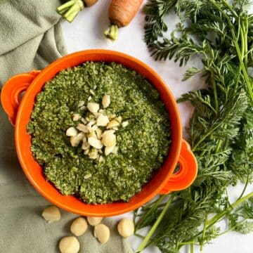 Orange bowl with carrot top pesto topped with chopped macadamia nuts and carrot tops on the side.
