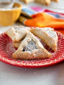 Chocolate-filled hamantaschen on a red plate with a yellow bowl in the background with powdered sugar suggesting the Mexican wedding cookie flavors as well.