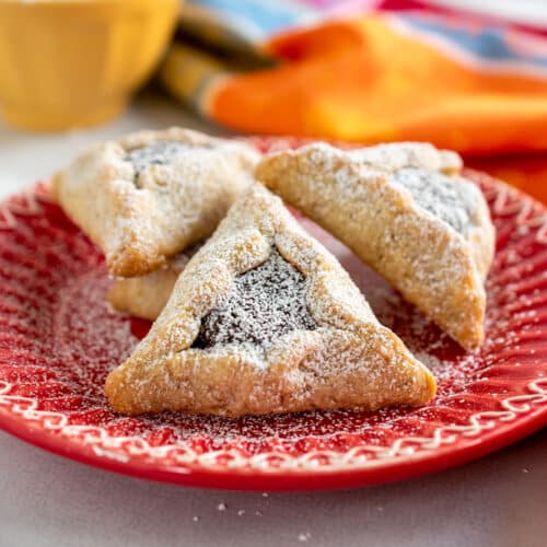 Chocolate-filled hamantaschen on a red plate with a yellow bowl in the background with powdered sugar suggesting the Mexican wedding cookie flavors as well.