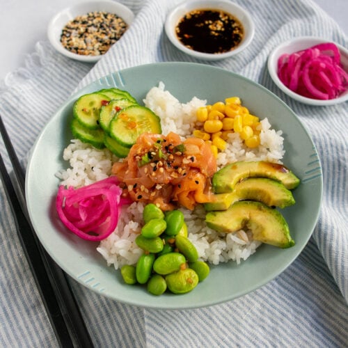 Smoked salmon poke bowl at an angle on a striped light blue napkin with black chopstics and 3 white bowls of seasonings in the back.