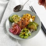 Hand sprinkling seasoning from a spoon onto a smoked salmon poke bow.