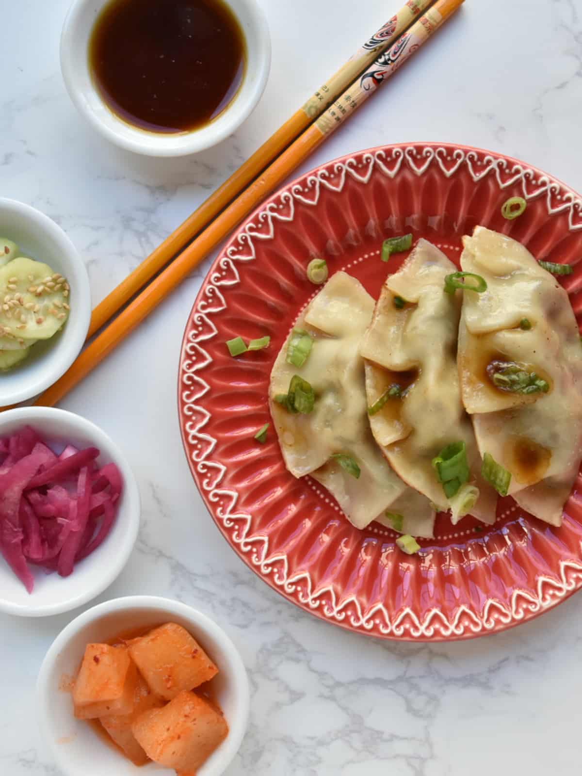 Jew Mandu (Korean kreplach) on a red plate with orange chopsticks and hapa banchan plus dipping sauce on the side.